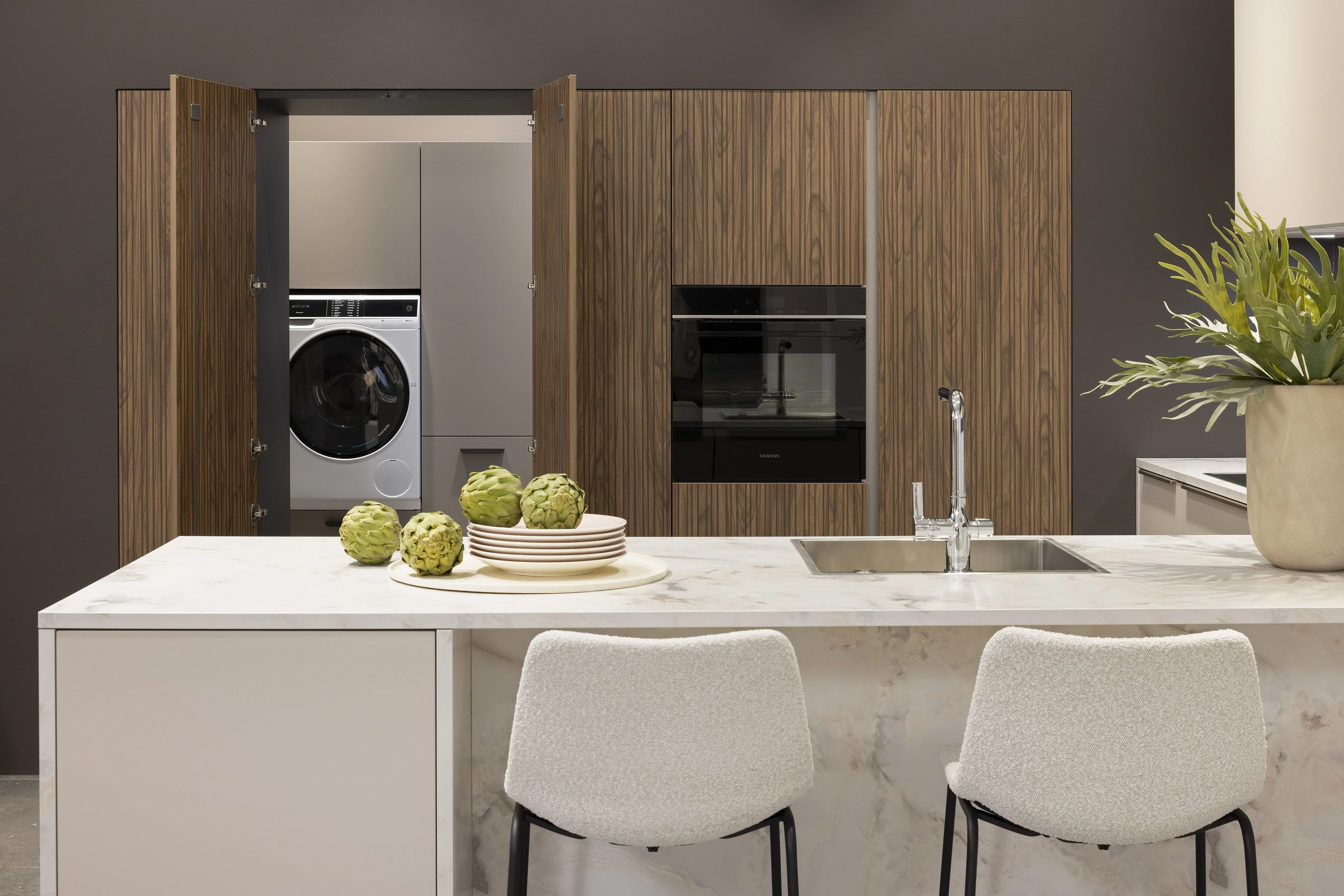 APL: Marmor Bianco 25 mm NB Front: Feel Sahara softmatt Front: WArtline Stripe Wood Choco Korpus: Sahara und Graphit Griff: Kehlleiste Edelstahloptik, und Griffe Edelstahloptik Geräte: Siemens  Armatur: Reginox Spüle: Reginox   Features: eingekoffertes Schrankelement, Tür zum HWR in Frontmaterial, niederer Sockel, Hängeoberschränke asymmetrisch mit Dunstabzug (als Esse geplant), Ecklösung mit LeMans Auszug, Theke mit Essplatz, Ordnungssystem Holz in Auszügen, Downlight in Arbeitsplatzbeleuchtung 